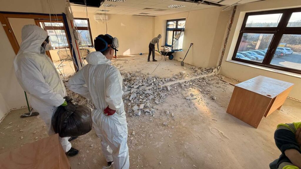 Budynek w trakcie prac demontażowych oraz pracownicy w strojach ochronnych wykorzystywanych podczas usuwania wełny ocieplającej strop